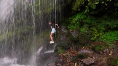 Günlük giysili genç bir kadın kayaların üzerinden tırmanır, canlı yeşillik ve doğal güzelliklerle çevrili verimli tropikal ormanlardaki şelaleye doğru yol alır.