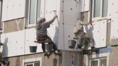 Asılı platformdaki inşaat işçileri dış duvar termal yalıtımını kaya yünü kullanarak kuruyorlar. Kask ve koşumlar da dahil olmak üzere güvenlik teçhizatı, yükseklikte güvenli çalışma koşullarını sağlar