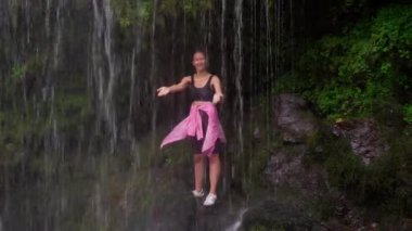 Güçlü şelalenin arkasında dikilen genç bayan turist gündelik giysiler giymiş ve mutlu bir şekilde gülümsüyor. Sahne, doğada seyahat ederken macera ve mutluluk hissi verir..