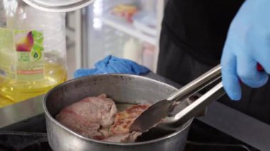 Mavi eldivenli aşçı tavadaki biftekleri katlamak, çevirmek ve düzenlemek için maşa kullanır. Restorandaki profesyonel mutfakta tava ocağın üzerinde duruyor..