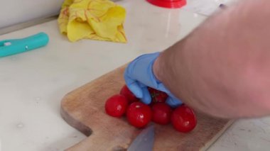 Mavi eldiven giyen aşçı, mutfaktaki ahşap kesme tahtasına kiraz domatesleri yerleştiriyor. Tezgahta patates ve bir tabak marul var.