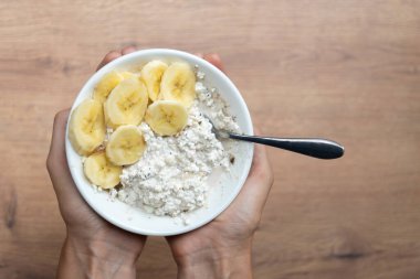 Kadın eli beyaz bir kase içinde süzme peynir, muz ve lifle ahşap bir masa tutar, sağlıklı yemek için idealdir..