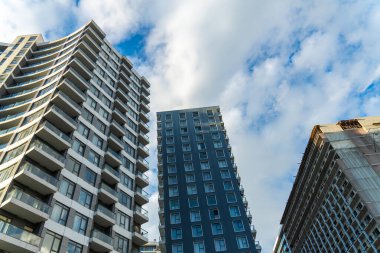 Modern apartman binaları, cam balkonlar, mavi gökyüzünün altında beyaz bulutlar, çağdaş şehir mimarisini gözler önüne seriyor.