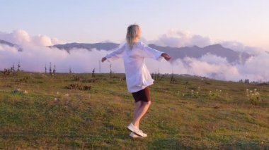 Sarışın, dağ manzarası ve bulutların çarpıcı güzelliğinin arka planında kolları açık mutlu bir şekilde döner. Doğa ile çevrelenmiş, huzurlu bir ortamda özgürlük ve mutluluk hissi yayar. Yavaş çekim