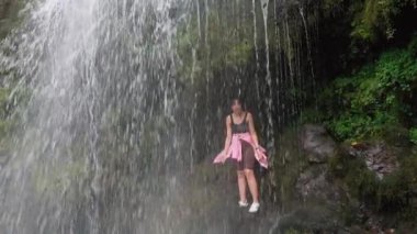 Güçlü şelalenin arkasında dikilen genç bayan turist gündelik giysiler giymiş ve mutlu bir şekilde gülümsüyor. Sahne, doğada seyahat ederken macera ve mutluluk hissi verir..