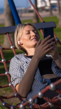 Happy blonde young woman in a plaid shirt lies in a hammock in the park with a smartphone and listens to music through white wireless headphones. Vertical photo clipart