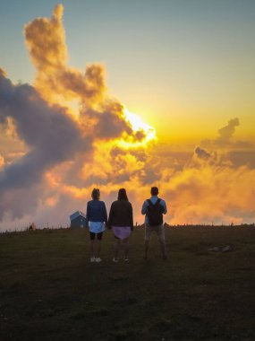 Three friends are enjoying the sunset at the top of the mountain, feeling freedom and happiness in nature, enjoying the picturesque beauty of standing above the clouds. Vertical photo clipart