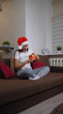 Genç bir kadın kahverengi koltukta bağdaş kurup kırmızı Noel hediye kutusunu açıyor. Noel Baba şapkası takıyor ve bayram ve sürpriz beklentisiyle gülümsüyor. dikey video