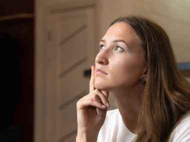 Genç bir kadın evdeki bir kanepede oturuyor, eliyle çenesine dokunuyor ve dikkatlice yukarı bakıyor, bir karar ya da sorun üzerinde düşünüyor.