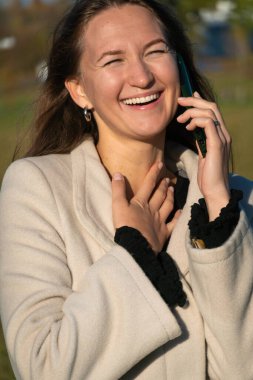 Güneşli bir günde, dışarıda telefonda konuşurken gülümseyen mutlu genç bir kadın, kolları bağlı, bej bir ceket giymiş ve bileklerinde fırfırlar olan siyah bir elbise giymiş. Dikey fotoğraf