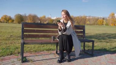 Autumn Park 'ta akıllı telefonuyla bankta oturan genç bir kadın. Sesli mesaj ya da video kaydı yaparken gülümsüyor ve gülüyor. Kahve içiyormuş. dikey video.