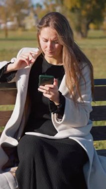 Parktaki bankta oturan genç bir kadın akıllı telefonundaki kısa mesajları okuyor, içeriği işlenmiş gibi görünüyor, hayal kırıklığı ve sinir bozukluğunu yansıtıyor.