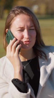 Parkta genç bir kadın, gülümsüyor ve telefonda konuşuyor. Neşeli tavırları, güneşli bir sonbahar gününde kendine güveni ve profesyonel yaşam tarzını yansıtıyor. dikey video