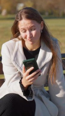 Parktaki bankta oturan bir kadın telefonla kitap okuyor ve dışarıda güneşleniyor. Sonbahar ağaçları dingin bir zemin oluşturur. dikey video