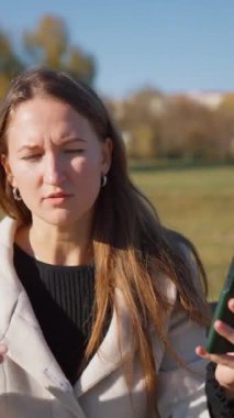 Parkta bankta oturan, gülümseyen ve telefonda konuşan genç bir kadın. Neşeli tavırları, güneşli bir sonbahar gününde kendine güveni ve profesyonel yaşam tarzını yansıtıyor. dikey video