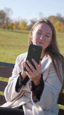 Sonbahar parkında akıllı telefon kullanan genç bir kadın. Sesli mesaj kaydederken ya da görüntülü konuşma yaparken gülümsüyor ve gülüyor. dikey video