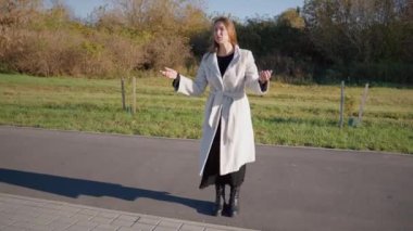 Beyaz önlüklü genç bir kadın arka planda çimenler ve ağaçlar olan asfalt bir yolda rol alıyor. Değişik duygular ifade ediyor ve işaret ediyor.