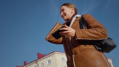 Genç bir kadın güneşli bir günde telefonuna bakarken endişe duyuyor. Sokakta dikiliyor ve bir şey hakkında çok endişeli görünüyor.