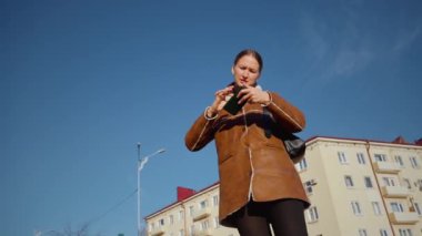 Genç bir kadın güneşli bir günde telefonuna bakarken endişe duyuyor. Sokakta dikiliyor ve bir şey hakkında çok endişeli görünüyor.