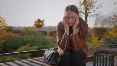 Parktaki bankta oturan iş kadını başını ellerinin arasına almış, baş ağrısıyla boğuşuyordu. Sonbahar doğasıyla çevrelenmiş, stresi ve yorgunluğu yansıtıyor.