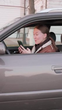 Araba süren genç bir kadın, heyecan verici haberler alınca şaşırdığını ve sevindiğini belirten akıllı telefonuna bakıyor. Onun mutlu tepkisi günlük hayatta iletişimin heyecanını vurguluyor. dikey video