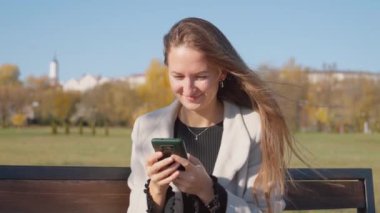 Parktaki bankta otururken telefonu kullanan genç ve güzel bir kadın. O, yanında uzun ceket ve bir bardak kahve giyer. Sonbahar Şehir Parkı.