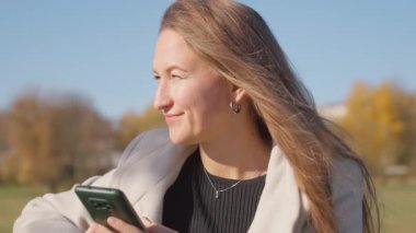 Parktaki bankta otururken telefonu kullanan genç ve güzel bir kadın. O, yanında uzun ceket ve bir bardak kahve giyer. Sonbahar Şehir Parkı.