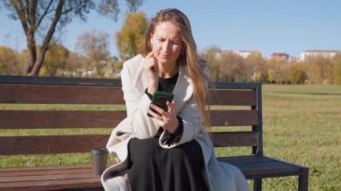 Parktaki bankta otururken telefonu kullanan genç ve güzel bir kadın. O, yanında uzun ceket ve bir bardak kahve giyer. Sonbahar Şehir Parkı.