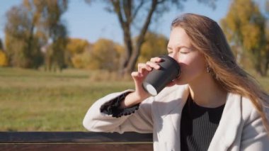 Genç bir kadın parkta bankta oturmuş, kağıt bardaktan sıcak içecek yudumluyor. Güneşli bir sonbahar gününde güneşin sıcaklığının tadını çıkararak doğanın huzurlu atmosferini kucaklıyor.