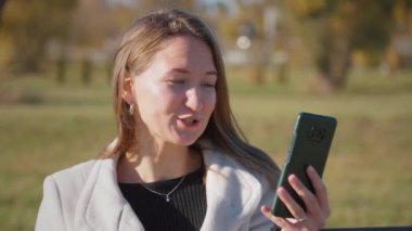Genç iş kadını ya da kız öğrenci bankta oturuyor, kahve içiyor ve video aracılığıyla telefonla konuşuyor. O, beyaz ceket ve siyah gömlek giyer. Neşeli gülümsüyor..
