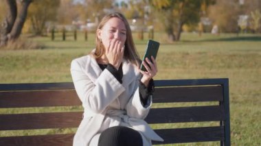 Genç iş kadını ya da kız öğrenci bankta oturuyor, kahve içiyor ve video aracılığıyla telefonla konuşuyor. O, beyaz ceket ve siyah gömlek giyer. Neşeli gülümsüyor..