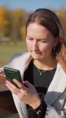 Parktaki bankta oturan genç bir kadın akıllı telefonuyla kısa mesaj okuyor, içeriğin işlendiği kadar kızgın, hayal kırıklığı ve sinir bozukluğunu yansıtıyor. Dikey video