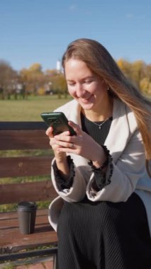 Parktaki bankta oturan, akıllı telefon kullanan genç bir kadın. Beyaz önlük ve siyah elbise giyerek, güneşli bir sonbahar gününden zevk alıyor, internete bağlanırken rahatlıyor ve gülümsüyor. dikey video