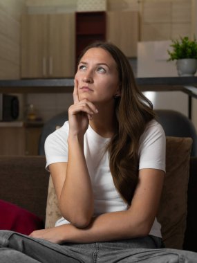 Genç bir kadın evdeki bir kanepede oturuyor, eliyle çenesine dokunuyor ve dikkatlice yukarı bakıyor, bir karar ya da sorun üzerinde düşünüyor. Dikey fotoğraf