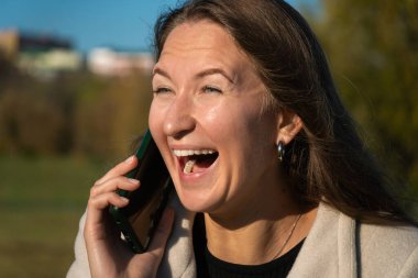 Güneşli bir günde, dışarıda telefonda konuşurken gülen mutlu genç bir kadın, kollarını kavuşturmuş, bej bir ceket giymiş ve bileklerinde fırfırlar olan siyah bir elbise giymiş.