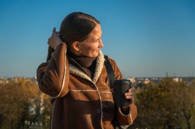 Kahverengi ceketli genç bir kadın köprünün parmaklıklarında oturuyor, elinde sıcak bir içecek olan bir kağıt bardak tutuyor ve sonbahar manzarasının tadını çıkarıyor.