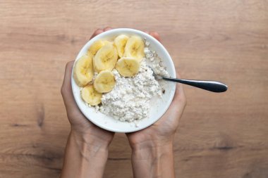 Kadın eli beyaz bir kase içinde süzme peynir, muz ve lifle ahşap bir masa tutar, sağlıklı yemek için idealdir..