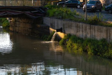 Su bir borudan nehre akıyor, doğayla karışıyor, endüstrinin çevre üzerindeki etkisini gösteriyor. Beton duvar, nehrin güzelliğiyle çelişiyor ve kirlilik sorununu vurguluyor