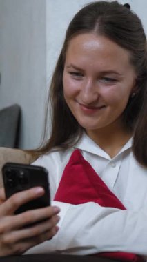 Genç kadın koltukta rahat bir şekilde oturuyor, arkasında kırmızı yastık, akıllı telefonunda kaydırıyor ve mesajlaşıyor, eğlenceden endişeye kadar çeşitli duygular sergiliyor.