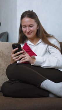 Genç kadın koltukta rahat bir şekilde oturuyor, arkasında kırmızı yastık, akıllı telefonunda kaydırıyor ve mesajlaşıyor, eğlenceden endişeye kadar çeşitli duygular sergiliyor.