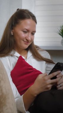 Genç kadın koltukta rahat bir şekilde oturuyor, arkasında kırmızı yastık, akıllı telefonunda kaydırıyor ve mesajlaşıyor, eğlenceden endişeye kadar çeşitli duygular sergiliyor.