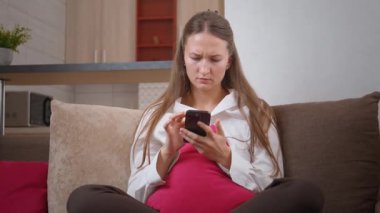 Genç kadın kanepeye oturur, akıllı telefon kullanır ve merakı, sıkıntısı, sürprizi ve hayal kırıklığı gibi duygular gösterir.