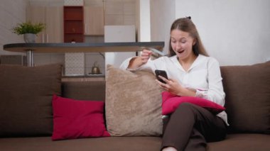 Kanepede oturan genç bir kadın akıllı telefonunu kullanarak iyi haberler okurken şaşkınlık ve mutluluk gösteriyor. Onun neşeli ifadesi sıcak ev ortamında heyecan ve neşeyi yansıtıyor.