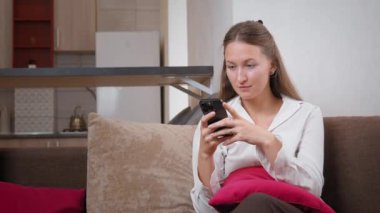 Kanepede oturan genç bir kadın akıllı telefonunu kullanarak iyi haberler okurken şaşkınlık ve mutluluk gösteriyor. Onun neşeli ifadesi sıcak ev ortamında heyecan ve neşeyi yansıtıyor.