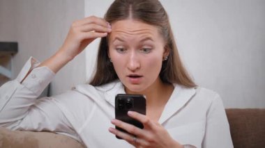 Kanepede oturan genç bir kadın akıllı telefonunu kullanarak iyi haberler okurken şaşkınlık ve mutluluk gösteriyor. Onun neşeli ifadesi sıcak ev ortamında heyecan ve neşeyi yansıtıyor.