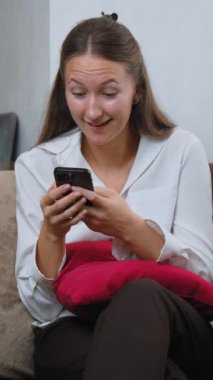 Kanepede oturan genç bir kadın akıllı telefonunu kullanarak iyi haberler okurken şaşkınlık ve mutluluk gösteriyor. Onun neşeli ifadesi sıcak ev ortamında heyecan ve neşeyi yansıtıyor.