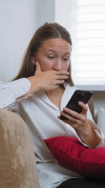 Kanepede oturan genç bir kadın akıllı telefonunu kullanarak iyi haberler okurken şaşkınlık ve mutluluk gösteriyor. Onun neşeli ifadesi sıcak ev ortamında heyecan ve neşeyi yansıtıyor.