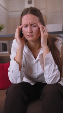 Kanepede oturan genç bir kadın çaresizlik içinde başını tutuyor, baş ağrısı çekiyor. Evdeki rahatsızlığıyla boğuşurken acı ve stres belirtileri gösteriyor. dikey video.