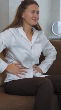 Kanepede oturan genç kadın şiddetli karın ağrısı çekiyor. Karnını tutuyor ve huzursuzluk içinde yüzünü buruşturuyor. dikey video