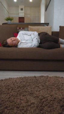 Kahverengi kanepede elini tutarak karın ağrısı çeken bir kadın. Şiddetli bir rahatsızlık yaşıyor, karnını tutuyor ve acı içinde kıvranıyor. dikey video